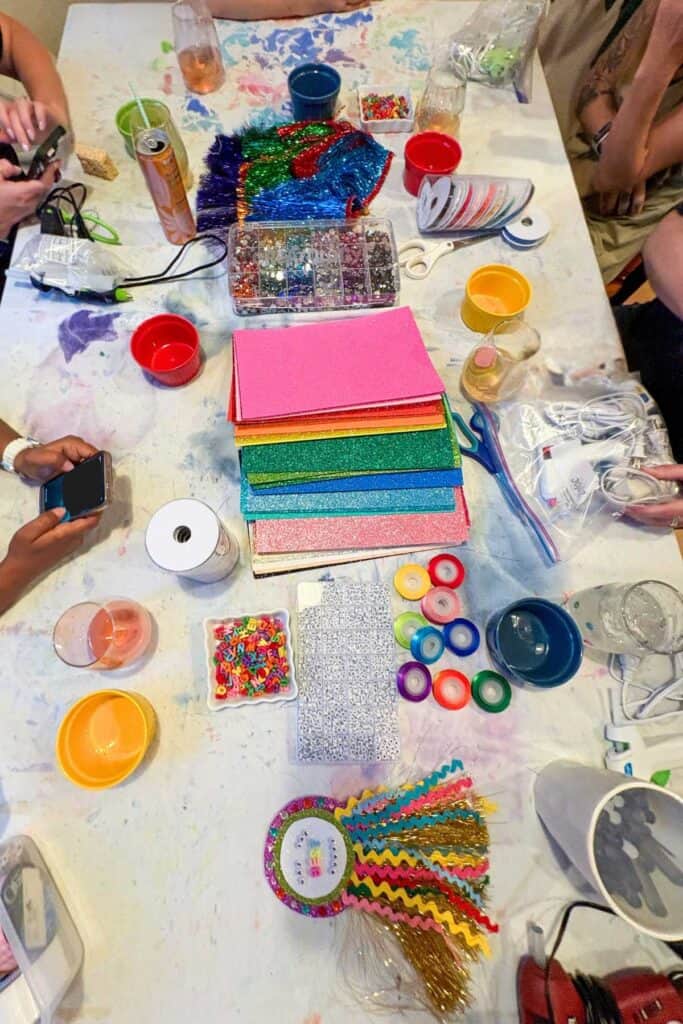 Craft night table covered in supplies for making DIY award badges including glitter foam sheets, rhinestones, letter beads, ribbon rolls, tinsel fringe, and hot glue guns with friends gathered around