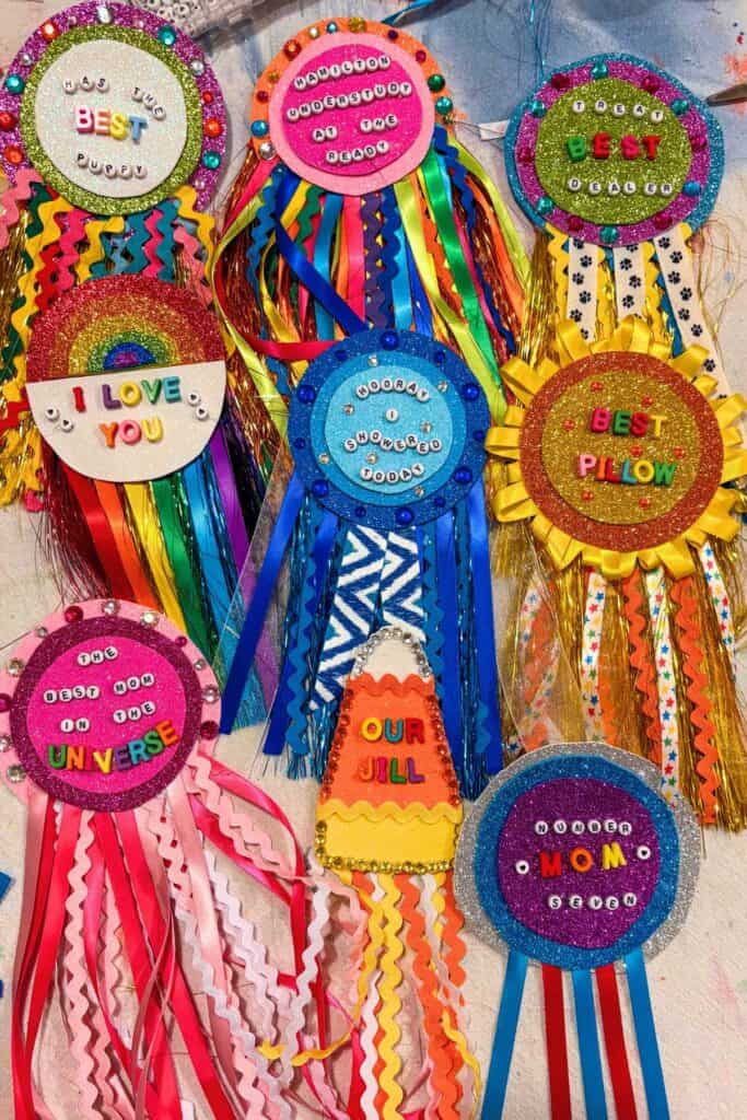 Flat lay of colorful handmade DIY award badges including "Hamilton Understudy at the Ready," "Best Pillow," "I Love You," "Hooray I Showered Today," and "The Best Mom in the Universe" made with glitter foam, letter beads, rhinestones, and ribbon fringe