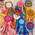 Collection of handmade DIY award badges laid out together including "Has the Best Puppy," "Hamilton Understudy at the Ready," "Treat Best Dealer," "I Love You" rainbow badge, "Hooray I Showered Today," "Best Pillow," "The Best Mom in the Universe," "Our Jill," and "Number Mom Seven" made with glitter foam, ribbon fringe, and letter beads
