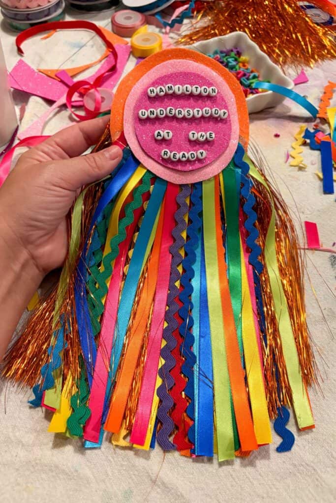 Close-up of a pink and orange glitter foam DIY award badge reading "Hamilton Understudy at the Ready" in white alphabet beads with an enormous colorful mixed ribbon and ric rac fringe tail in every color of the rainbow