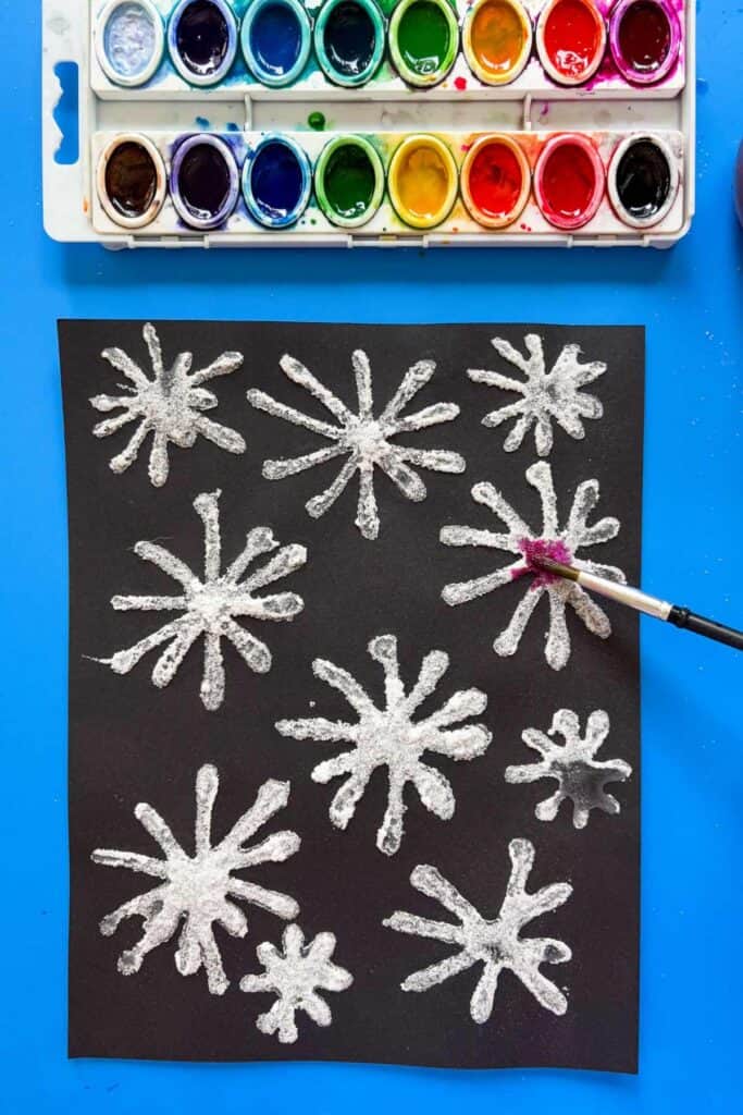 A paintbrush loaded with magenta watercolor is being tapped onto a black piece of paper covered in salt firework shapes, with a full watercolor palette visible at the top.