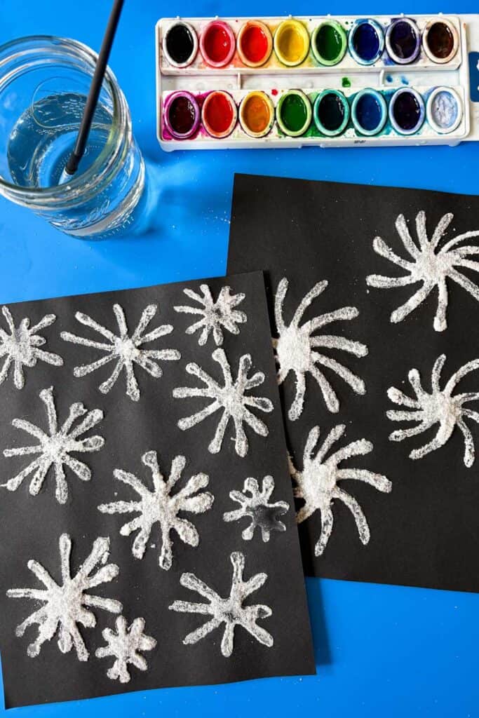 Two sheets of black paper covered in dried salt firework shapes lay on a blue surface next to a jar of water, a paintbrush, and a watercolor palette, ready for painting.