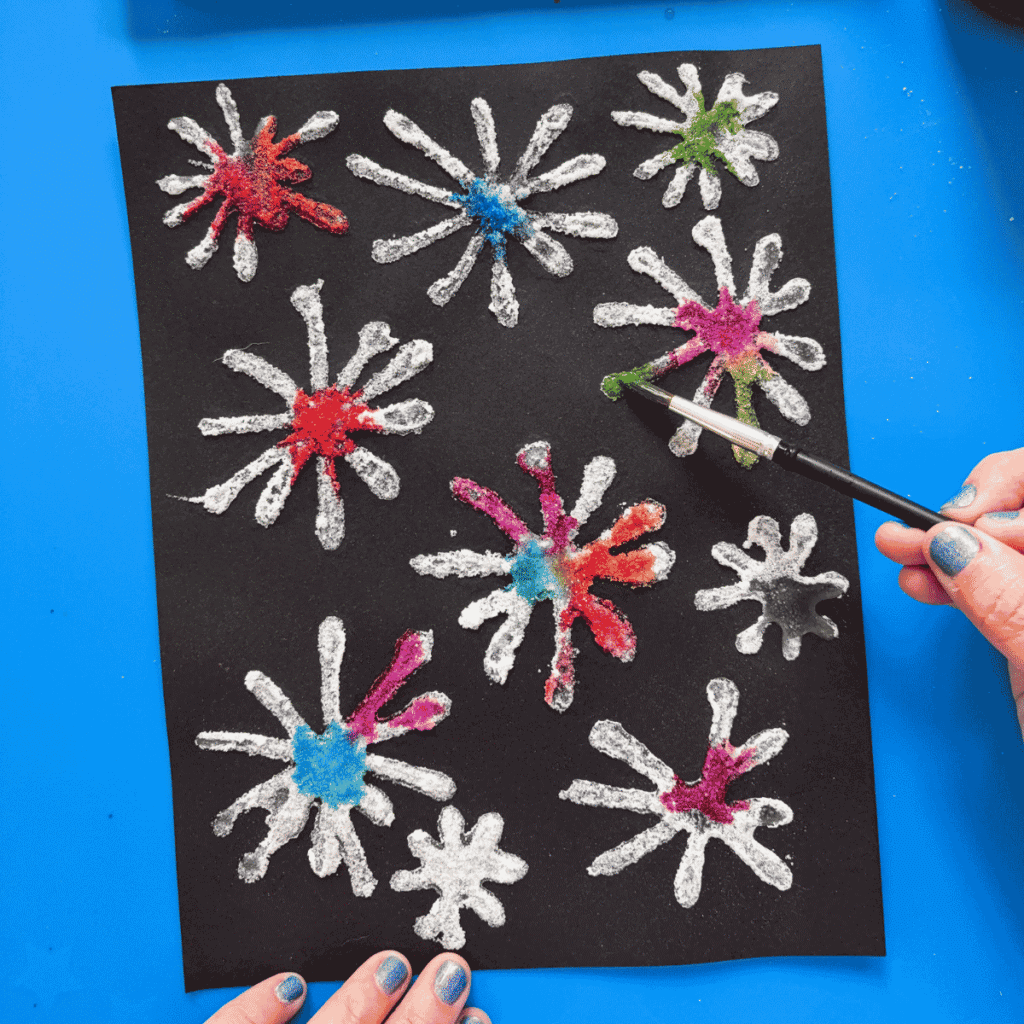 Completed Fourth of July craft salt firework art on black paper showing vibrant bursts of rainbow watercolor spread across starburst-shaped salt designs, held up against a blue background with a watercolor palette visible above.