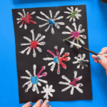 Completed Fourth of July craft salt firework art on black paper showing vibrant bursts of rainbow watercolor spread across starburst-shaped salt designs, held up against a blue background with a watercolor palette visible above.