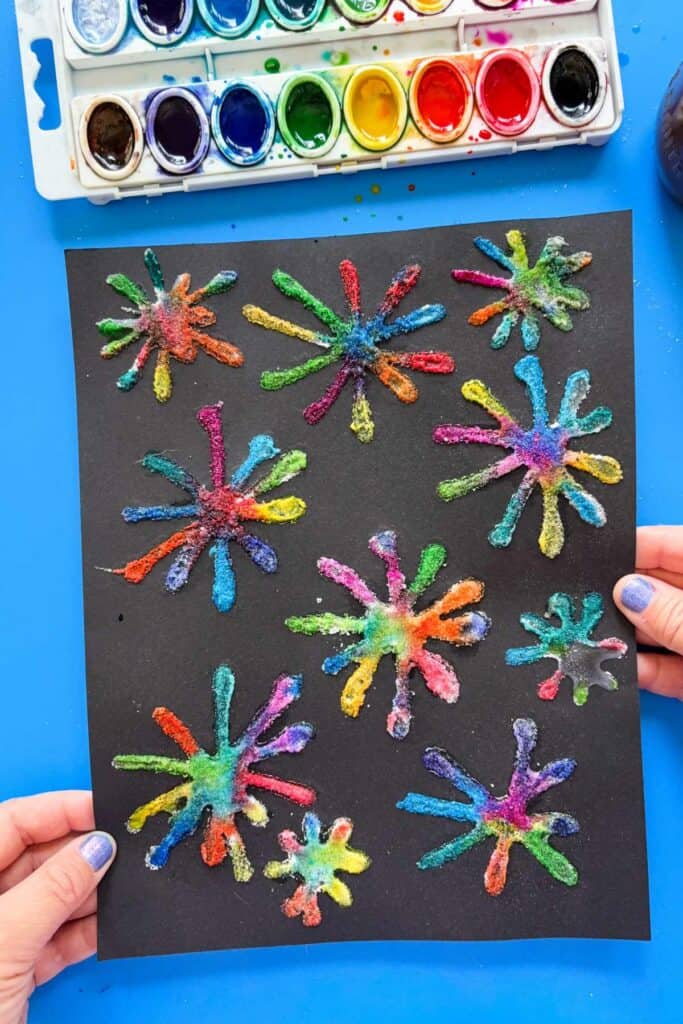 Two hands hold up a finished sheet of black paper filled with colorful salt fireworks in every color of the rainbow, with a watercolor palette visible in the upper corner.