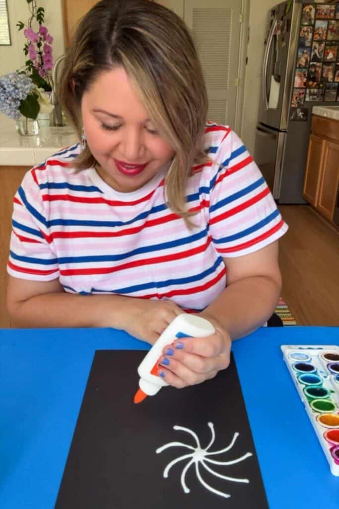 A woman in a red, white, and blue striped shirt smiles as she squeezes white school glue onto black paper to draw a firework shape, with a watercolor palette set to the side, for the Fourth of July craft.