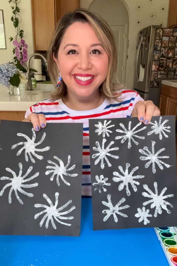 A smiling woman in a red, white, and blue striped shirt holds up two sheets of black paper covered in dried salt firework shapes, ready to be painted with watercolors.