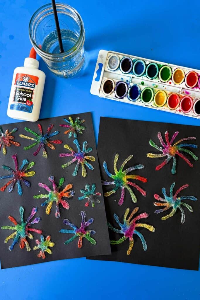 Flat lay of supplies needed for the Fourth of July craft salt fireworks: a bottle of Elmer's school glue, a jar of water with a paintbrush, a watercolor palette, and two completed sheets of colorful salt firework art on black paper, all arranged on a bright blue surface.