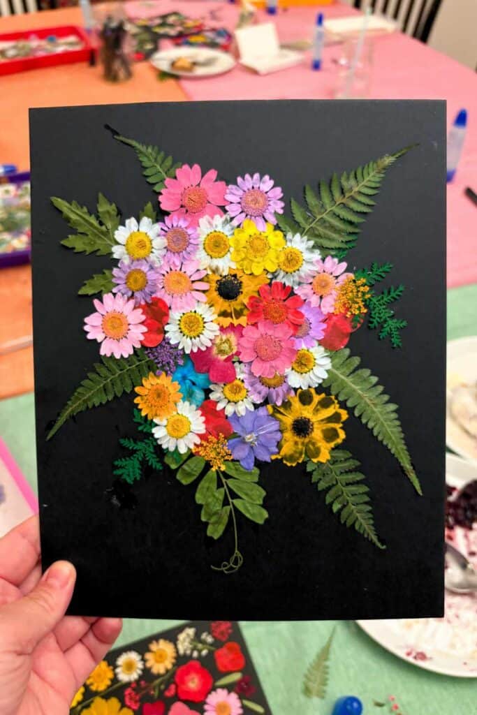 A hand holds up a pressed flower art piece on black mat board with a lush bouquet-style arrangement of colorful dried flowers surrounded by large fern fronds and greenery spread across the corners, with a craft night table visible in the background.