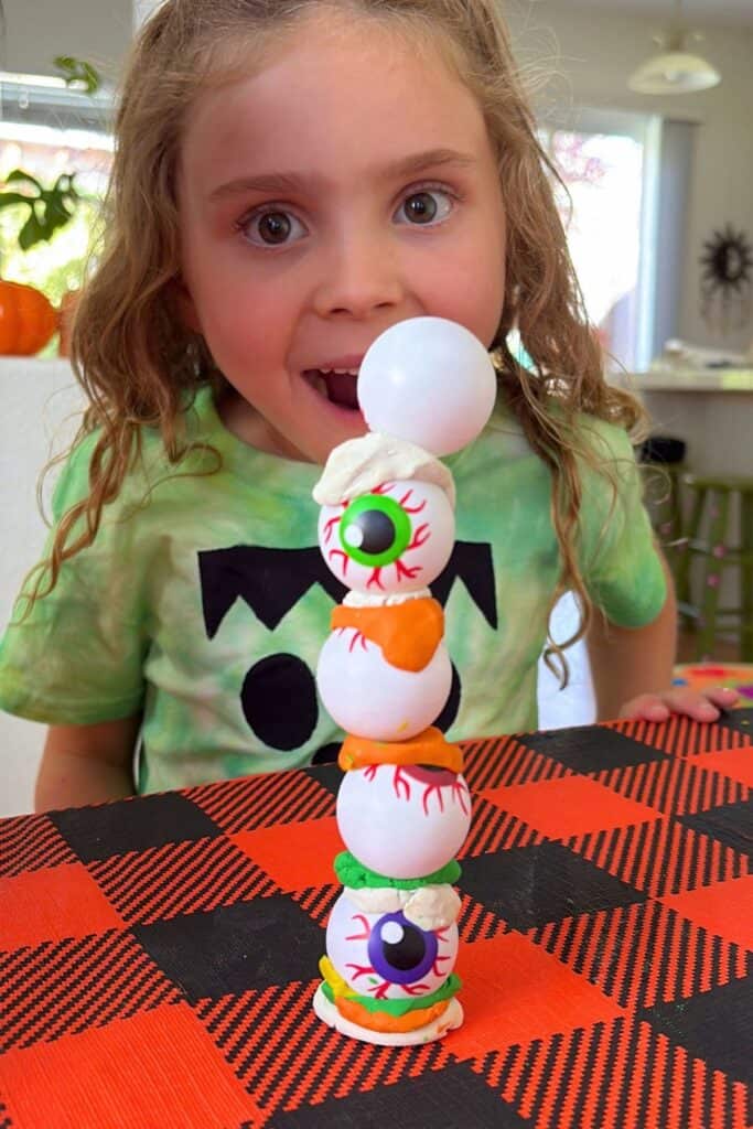 A young girl gasps in delight next to her five-ball eyeball tower, part of this Halloween STEM activity, with a bite mark clay layer at the top.