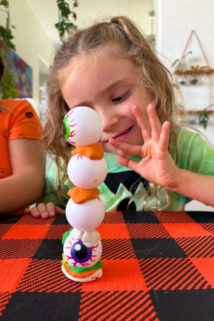 A girl carefully lets go of her four-ball eyeball tower to test whether the clay layers can hold it steady on their own.