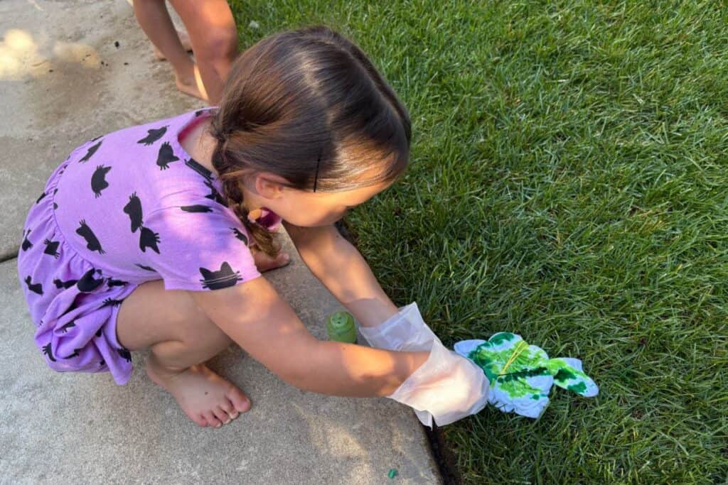 A child applying green dye to a rubber-banded shirt spiral, kneeling on the grass during the tie-dye process.