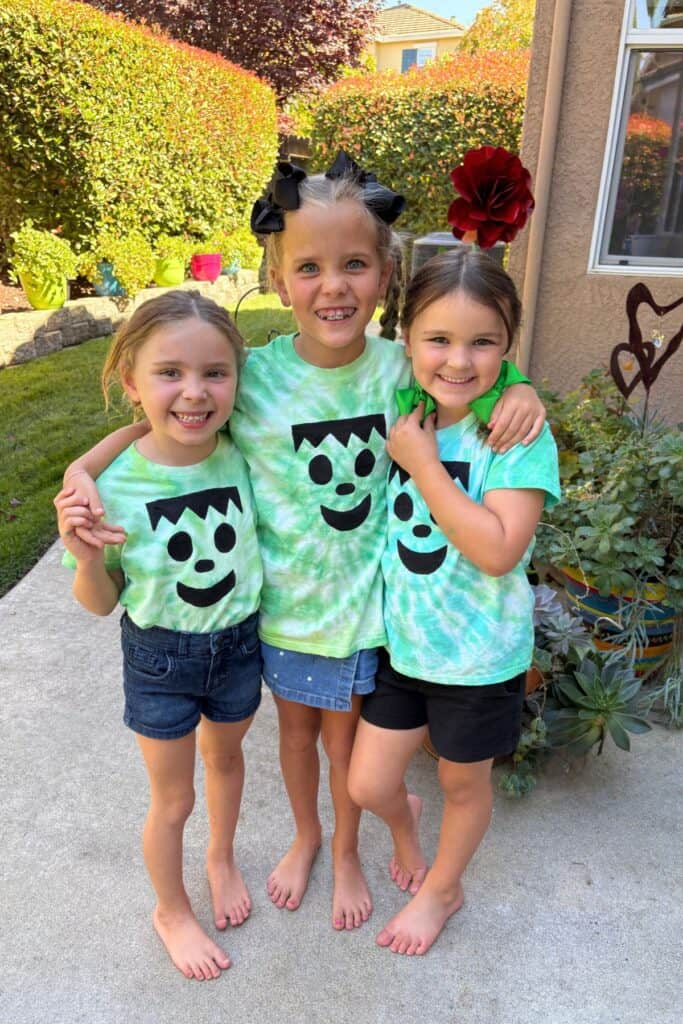 Three kids smiling together outdoors in their Frankenstein Halloween tie-dye shirts, each with a different green swirl pattern and painted monster face.