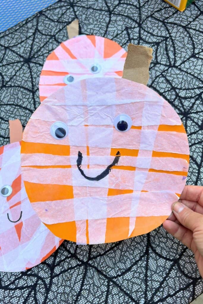 Child holding a finished jack-o-lantern craft for Halloween decorated with layered white tissue paper and googly eyes.
