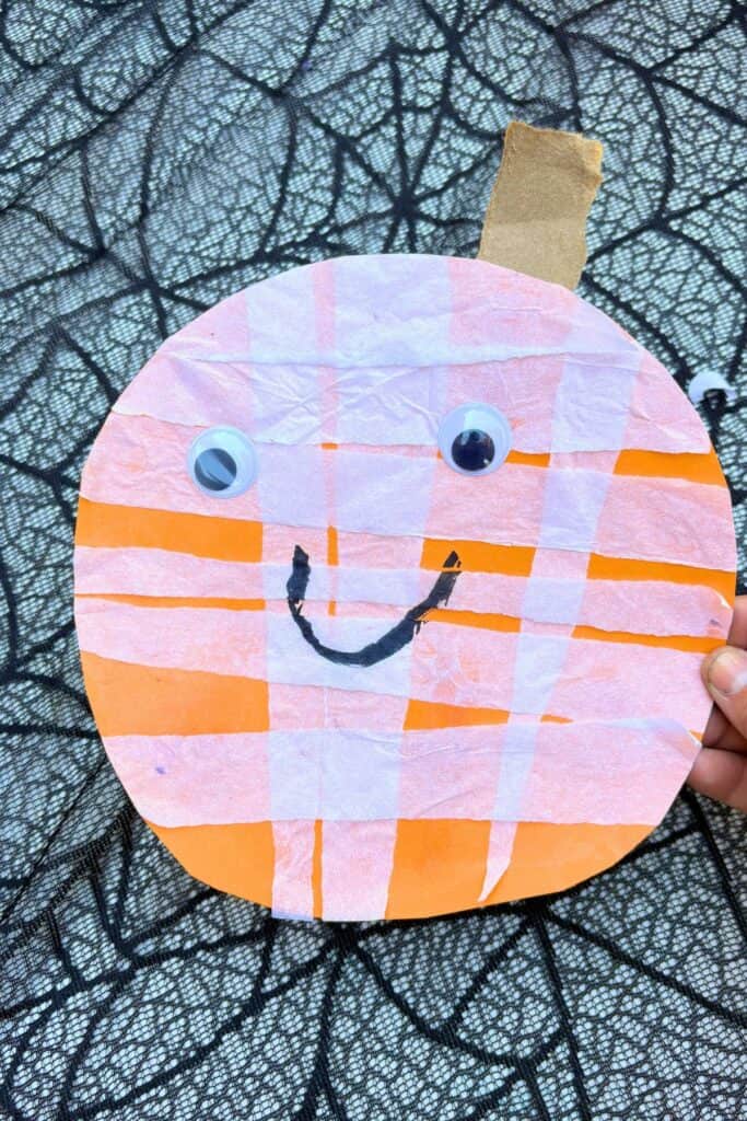 Jack-o-lantern craft for Halloween made with orange paper, white tissue paper strips, googly eyes, and a hand-drawn smile.