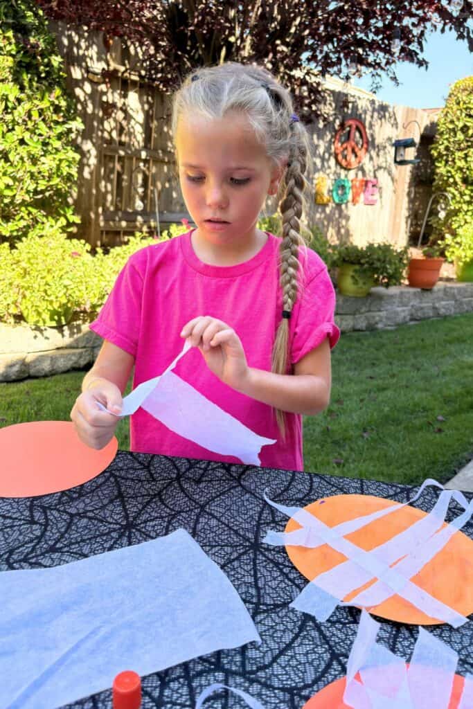 Child tearing white tissue paper into strips while making a jack-o-lantern craft for Halloween at an outdoor craft table.