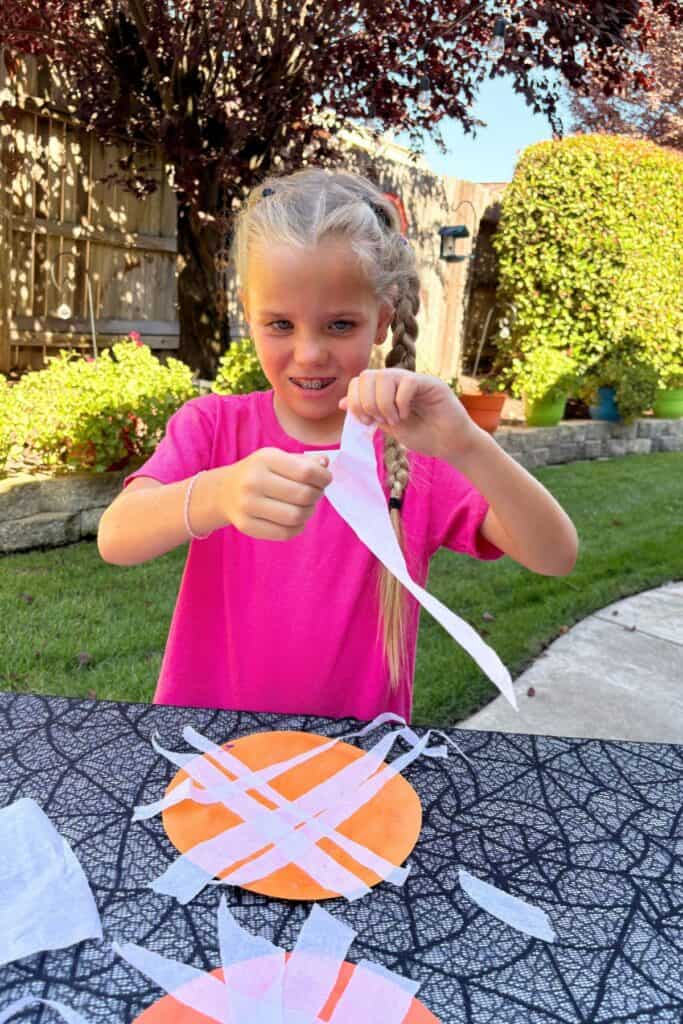 Smiling child tearing long strips of white tissue paper for a mummy pumpkin Halloween craft.