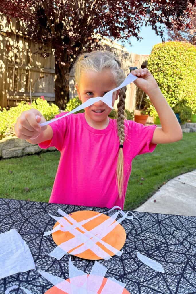 Child stretching and tearing white tissue paper above orange paper pumpkins during a Halloween craft activity.
