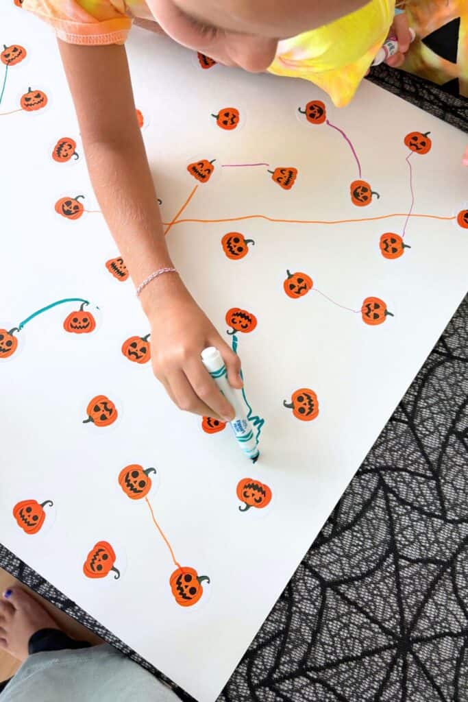 Child using a teal marker to connect matching pumpkin stickers during a preschool Halloween activity.