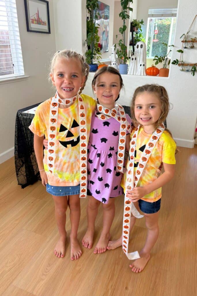 Three smiling children wearing long strands of jack-o-lantern stickers after completing a Halloween matching game.