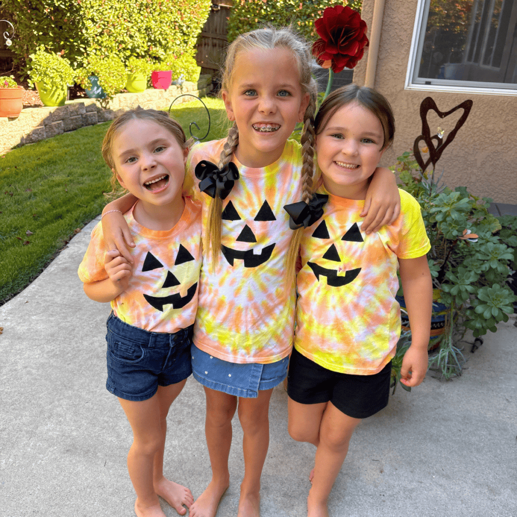 Three kids show off their finished jack-o-lantern Halloween tie-dye shirts, each with a hand-painted pumpkin face in a different orange and yellow swirl pattern.