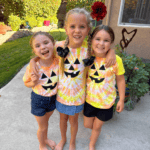 Three kids show off their finished jack-o-lantern Halloween tie-dye shirts, each with a hand-painted pumpkin face in a different orange and yellow swirl pattern.