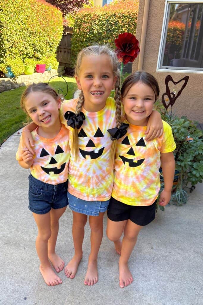 Three girls smiling and posing together in matching jack-o-lantern Halloween tie-dye shirts in the backyard.