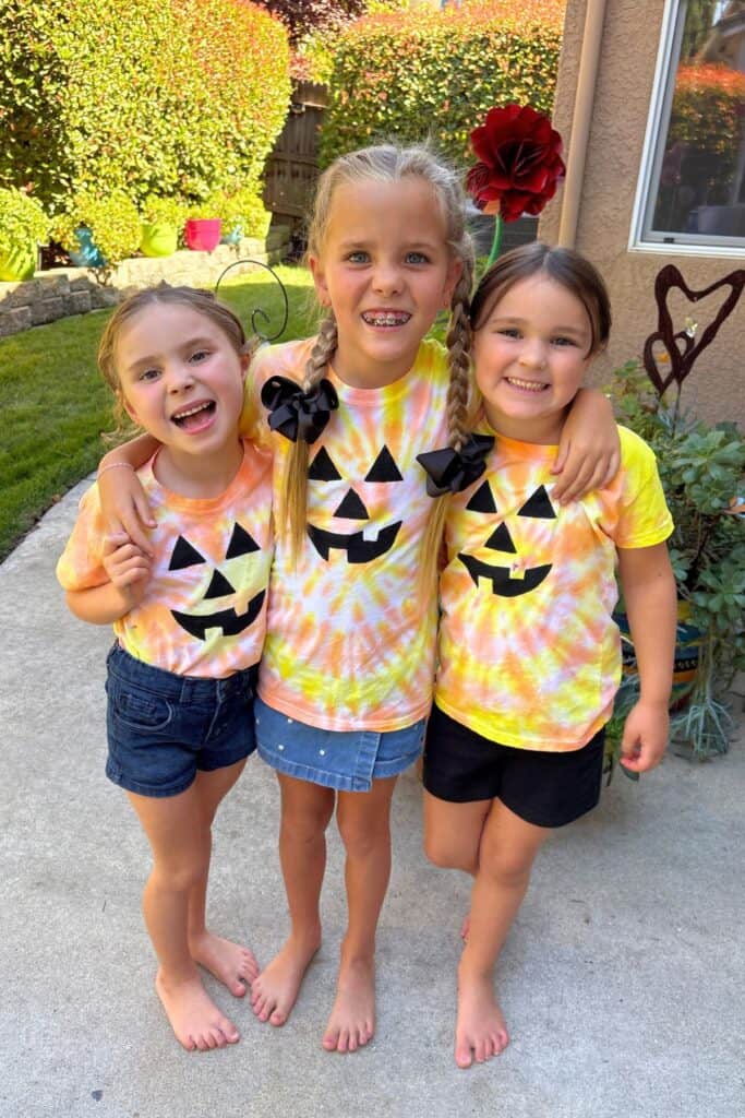 Three kids stand arm in arm outdoors, each wearing a jack-o-lantern Halloween tie-dye shirt with a black painted pumpkin face.